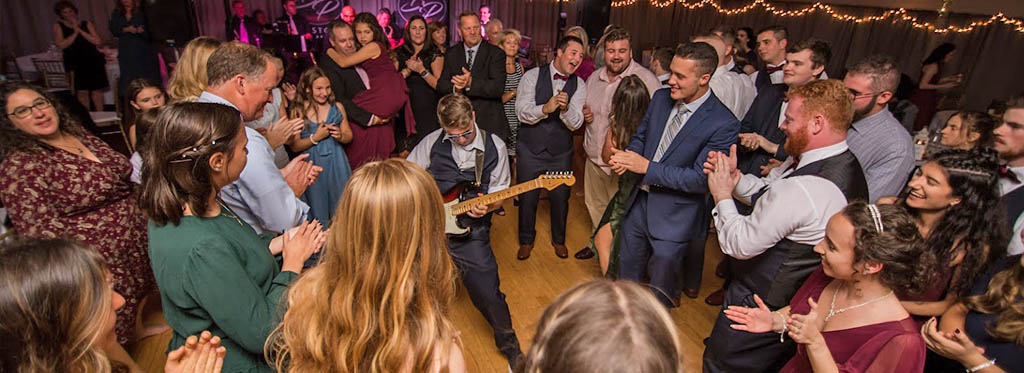 A guitarist from a Boston wedding band plays in the center of the dance floor with wedding guests surrounding him as he plays a solo.