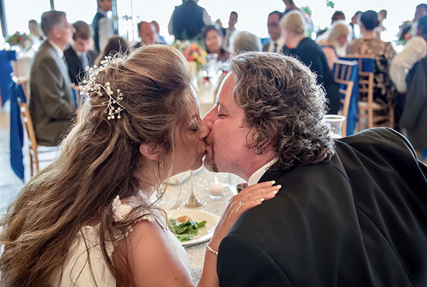 A wedding couple kiss at their table while guests applaud