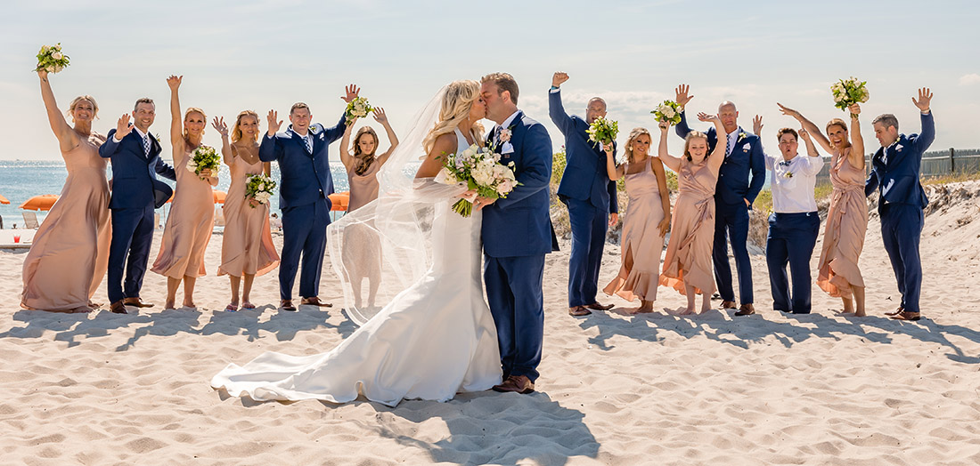 A bride and groom kissing in front of their bridal party on the beach.